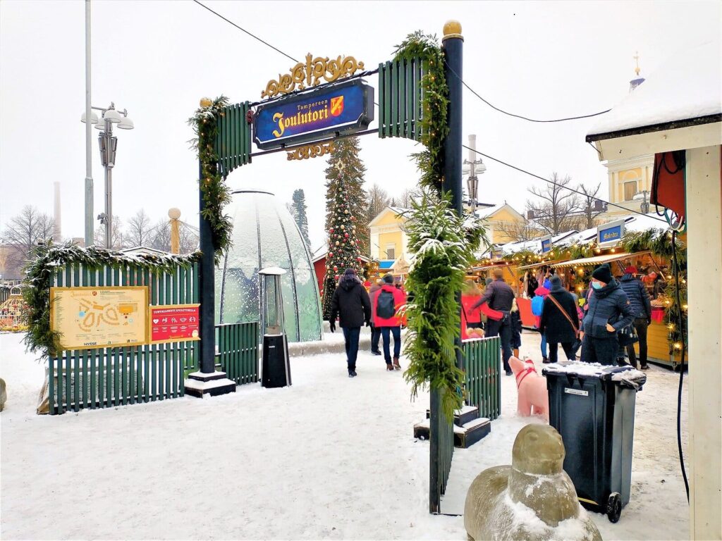 L’entrée du Joulutori, le marché de Noël de Tampere, sous une arche de verdure et de lumières, avec des chalets enneigés et la verrière du jet d’eau au fond