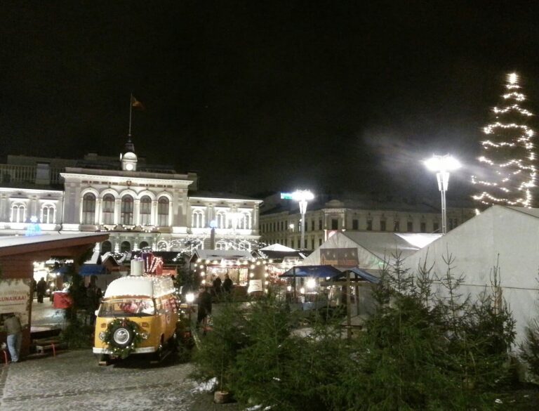 Le marché de Noël de la place centrale de Tampere de nuit, ses chalets et un petit camion de vente, avec l’ancien hôtel de ville illuminé au fond