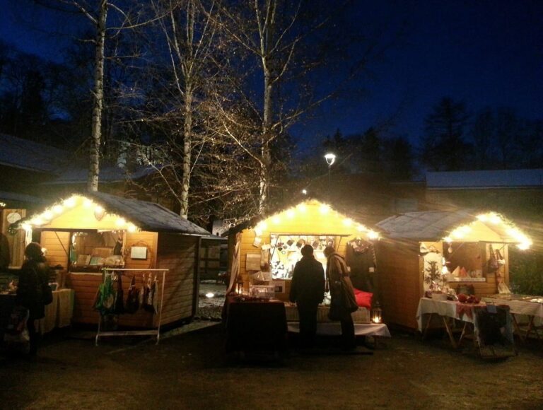 Des chalets de Noël éclairés dans la cour Tallipiha de Tampere, à la tombée de la nuit, entre des bouleaux