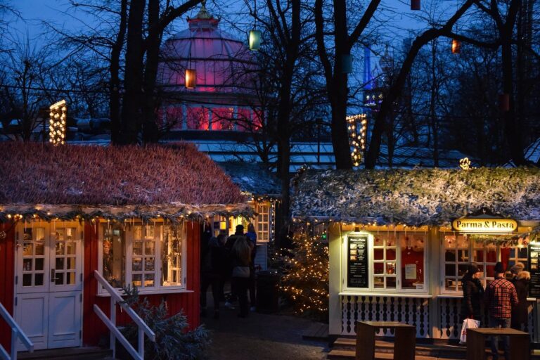 Deux chalets de bois aux toits givrés et décorés de guirlandes, avec le dôme éclairé en rose du Palais de verre derrière, dans les jardins de Tivoli