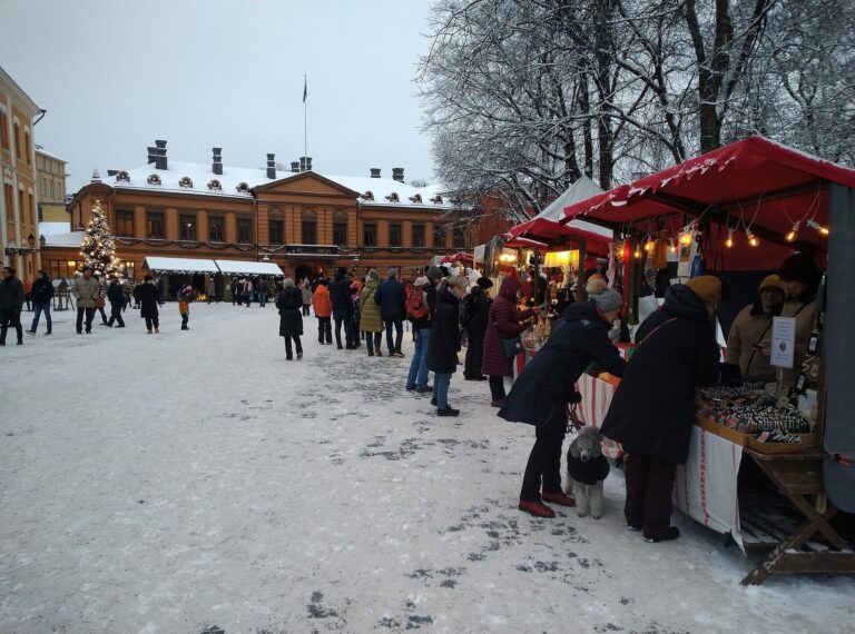 Des visiteurs devant un chalet du marché de Noël de la Vieille Grand-Place de Turku, sapin illuminé et maison Brinkkala au fond, sous la neige