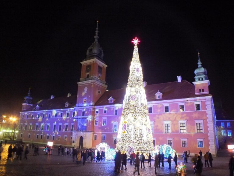 Le château royal de Varsovie éclairé en rose et en bleu, avec un grand sapin blanc illuminé sur la place, de nuit