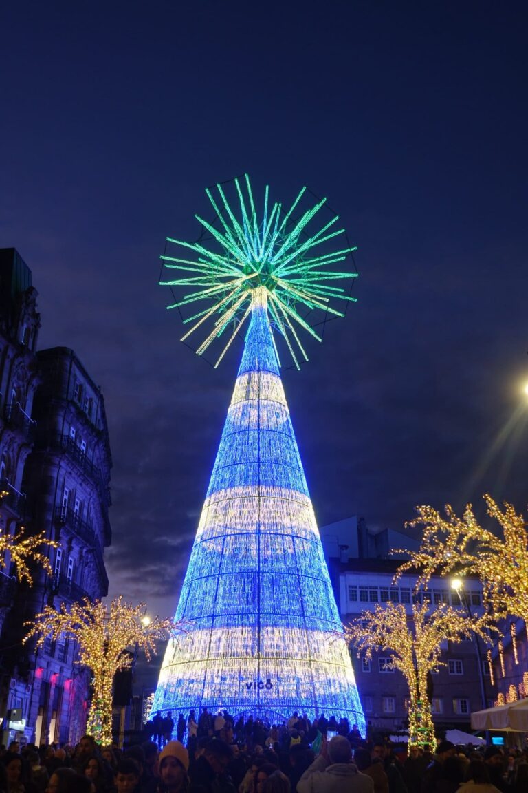 Le grand sapin lumineux de Vigo, cône bleu et blanc surmonté d’une gerbe de lumières vertes, entre des arbres illuminés à la tombée de la nuit