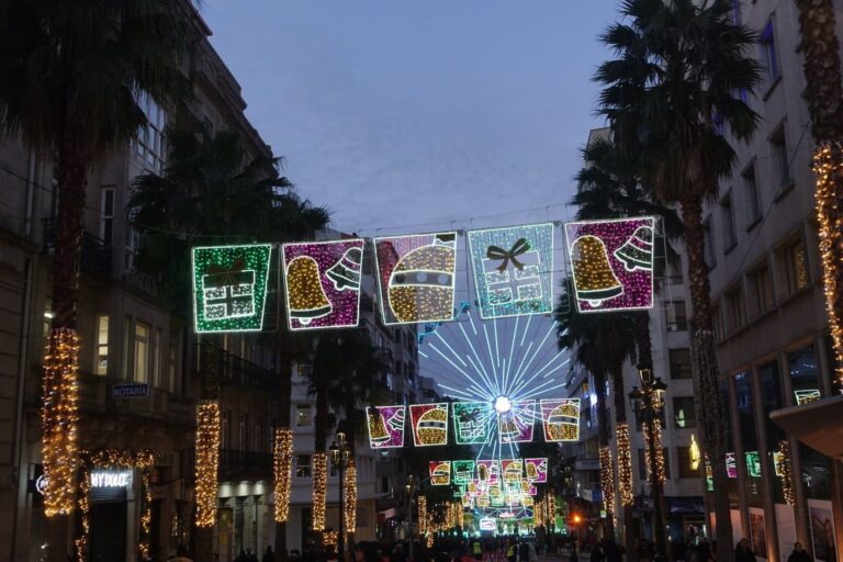 Une rue du centre de Vigo bordée de palmiers, sous des panneaux lumineux en forme de cadeaux, de cloches et de boules, avec la grande roue au fond, à la nuit tombante