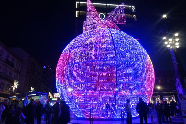 Une boule de Noël géante en guirlandes roses et bleues, surmontée d’un nœud, sur une place de Vigo, de nuit, avec des promeneurs vus de dos